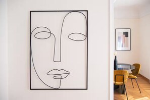 Une œuvre d’art minimaliste accrochée au mur, un coin repas avec une table ronde et des chaises jaunes, et un tableau encadré au mur.
