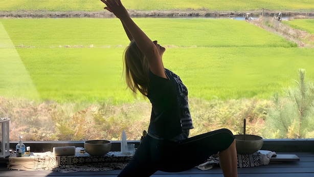 Una persona realiza una postura de yoga sobre una esterilla bajo una estructura de madera con vistas a un campo verde.