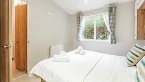 A bedroom with a bed, a window with curtains, a radiator, and a wooden door. - Sea Squirrel - Kelling Heath Holiday Park - 15856 (Holt)