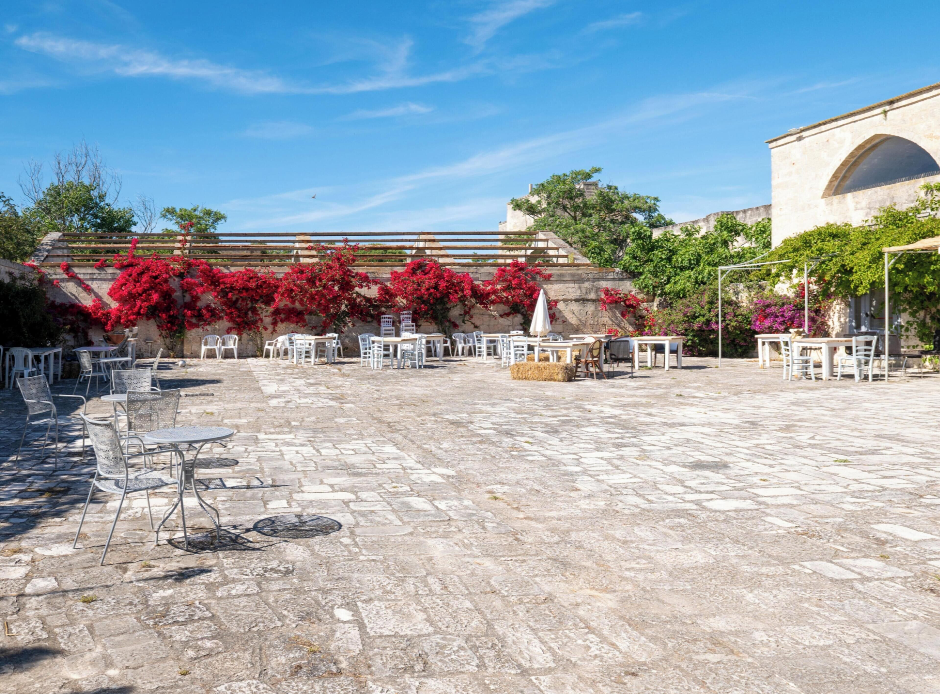 Un patio al aire libre con suelo de piedra, mesas y sillas blancas, y plantas con flores rojas.
