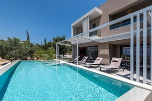 A modern poolside area with lounge chairs and a pergola, surrounded by a building with large windows and balconies. - Anthemis Luxury Villas 1 (Heraklion)