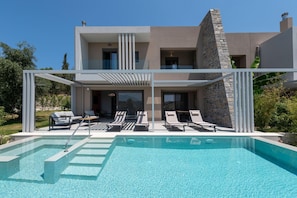 A modern two-story house with a swimming pool, lounge chairs, and a stone facade. - Anthemis Luxury Villas 1 (Heraklion)