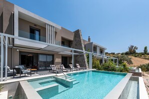 A modern poolside area with lounge chairs and a clear blue pool, surrounded by a contemporary building with a stone accent wall. - Anthemis Luxury Villas 1 (Heraklion)