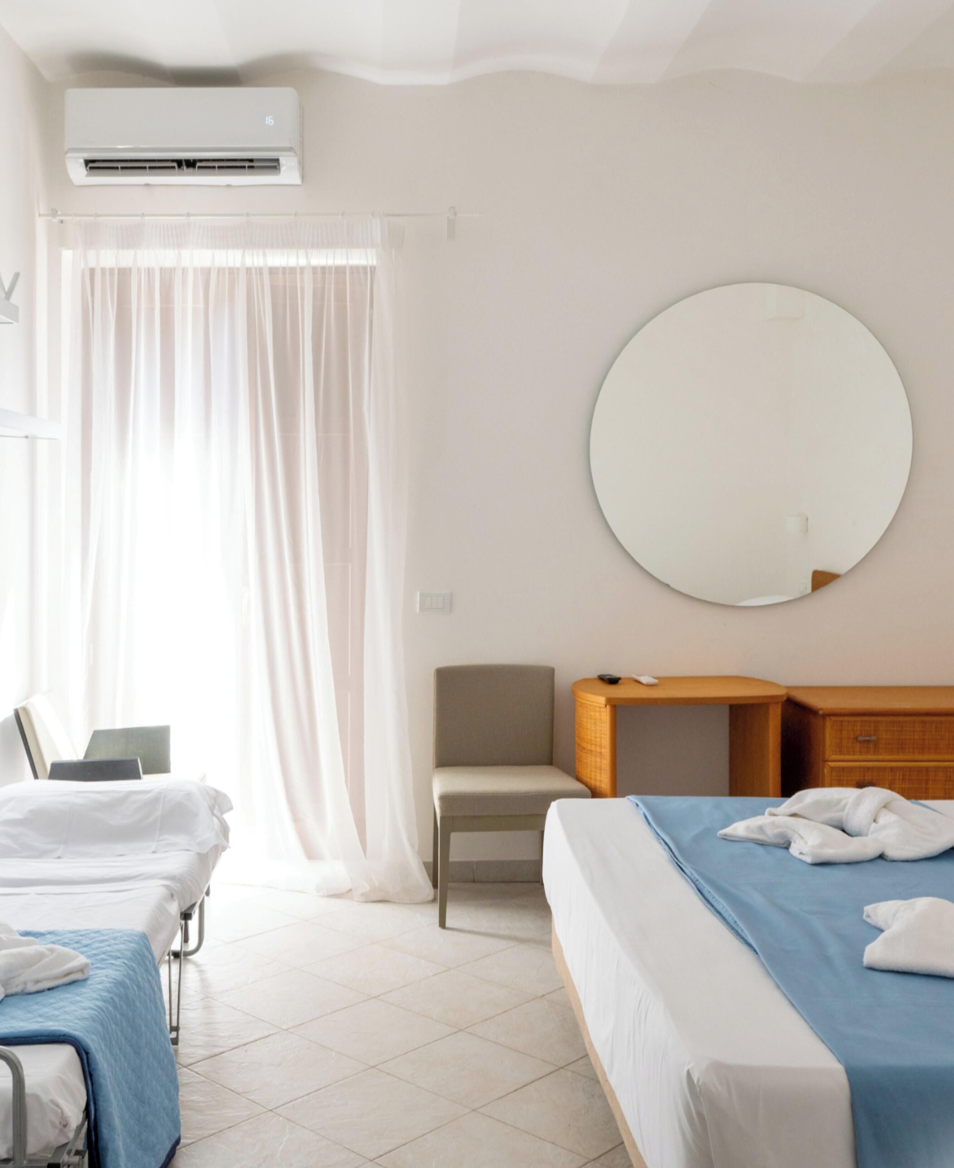 Une chambre d’hôtel avec deux lits, un miroir rond, une chaise, un bureau et un appareil de climatisation.