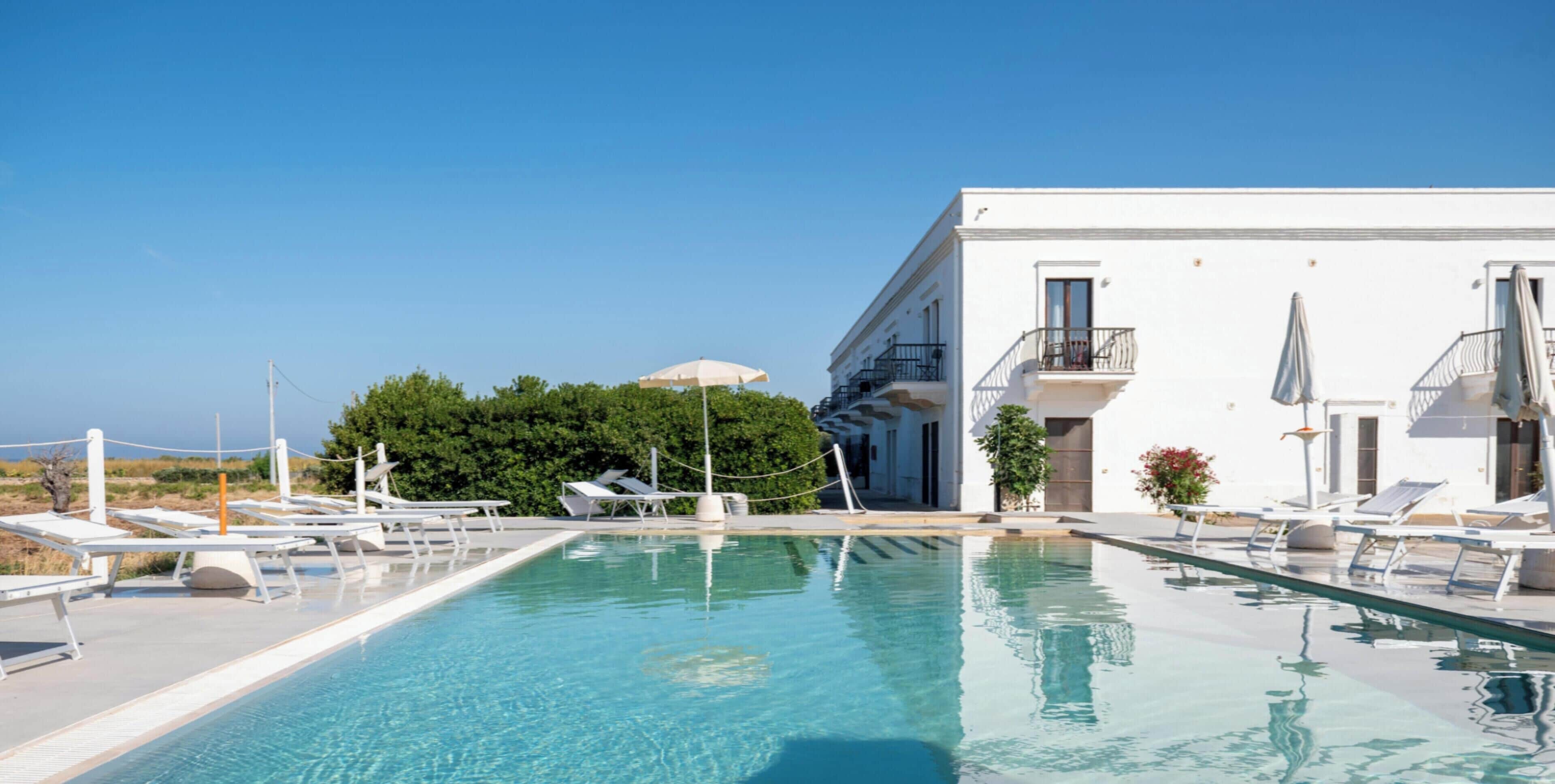 Une piscine avec des chaises longues et des parasols, un bâtiment blanc doté de balcons, et un ciel d’un bleu limpide.