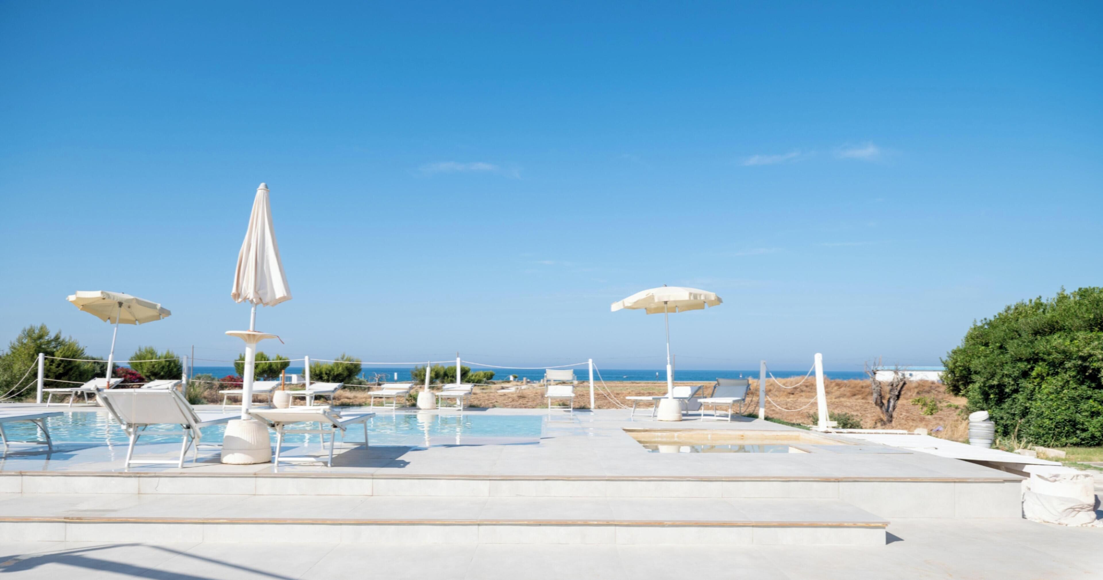 A pool area with lounge chairs and umbrellas, a clear sky, and a view of the sea.