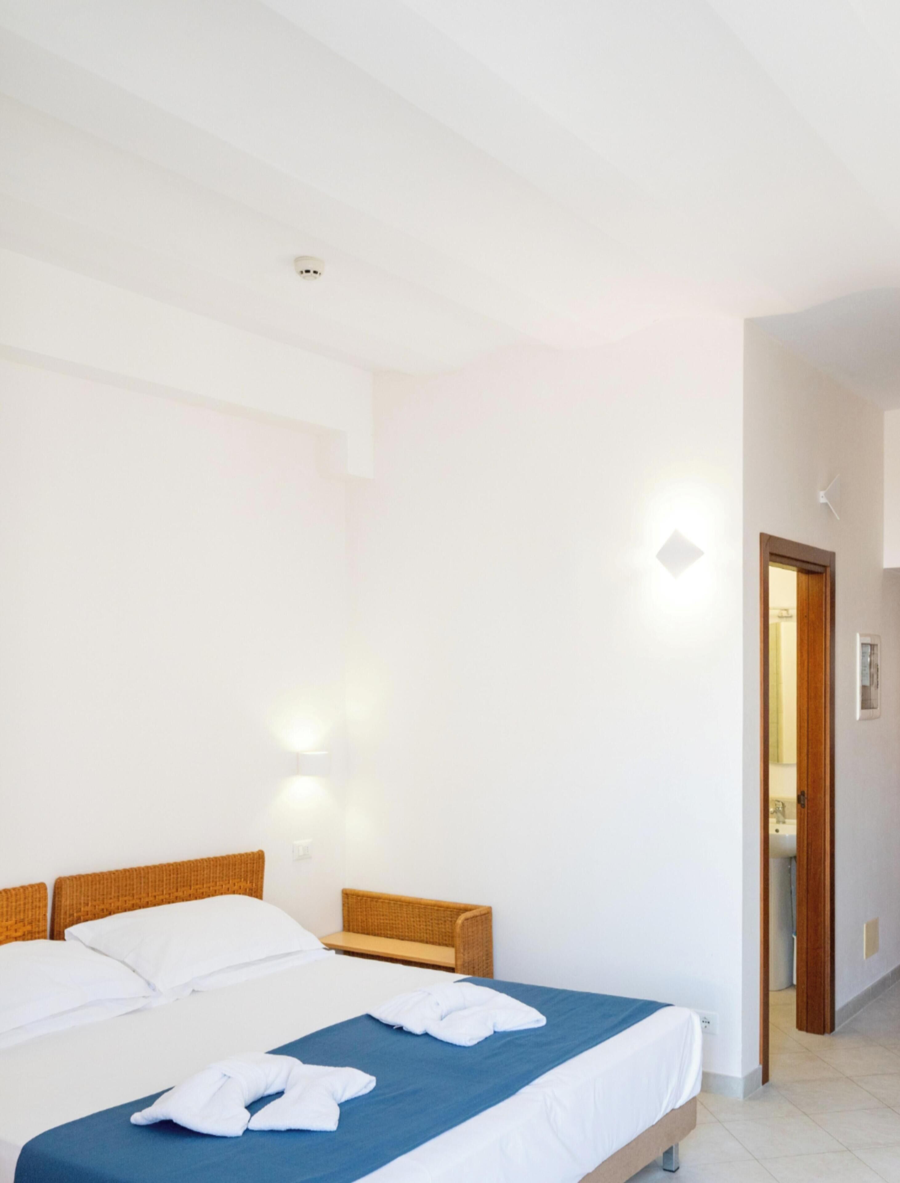 Hotelkamer met een tweepersoonsbed, een houten hoofdeinde, een blauwe spreidek en een witte muur met een lichtschakelaar.