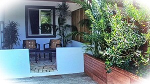 Una casa con facciata bianca, un patio in legno, un piccolo spazio esterno con tavolo e sedie, e un vaso grande con diverse piante.