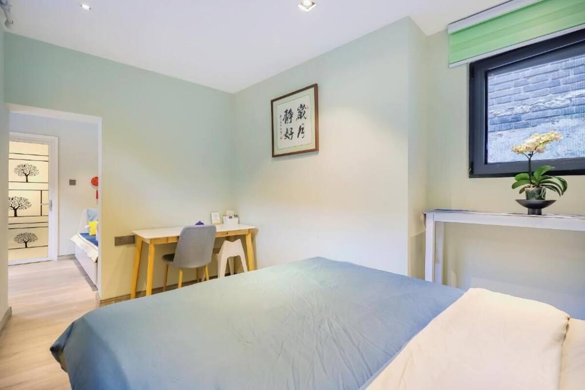 A bedroom with a bed, a desk, a chair, a window with a view, and a framed picture on the wall.