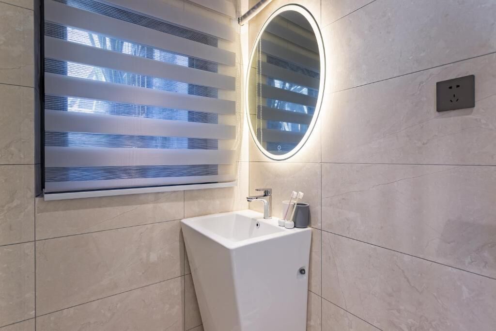 Un baño moderno con un lavabo blanco, un espejo y una ventana con persianas horizontales.
