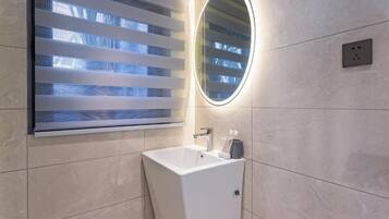 Un baño moderno con un lavabo blanco, un espejo y una ventana con persianas horizontales.
