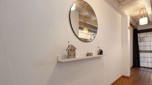 Un corridoio con una parete bianca, un pavimento in legno, uno scaffale bianco con un modellino di una casa e uno specchio rotondo.