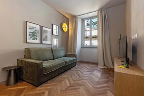 Ein modernes Wohnzimmer mit Fischgrätparkettboden, einem grünen Sofa, einem kleinen runden Tisch und einem Flachbildfernseher auf einem Holzgestell.