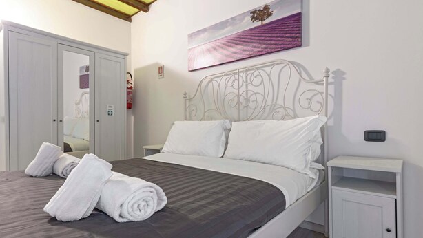 Un lit bien fait, avec du linge de lit blanc, une tête de lit en métal blanche et une table de chevet dotée d’un tiroir.