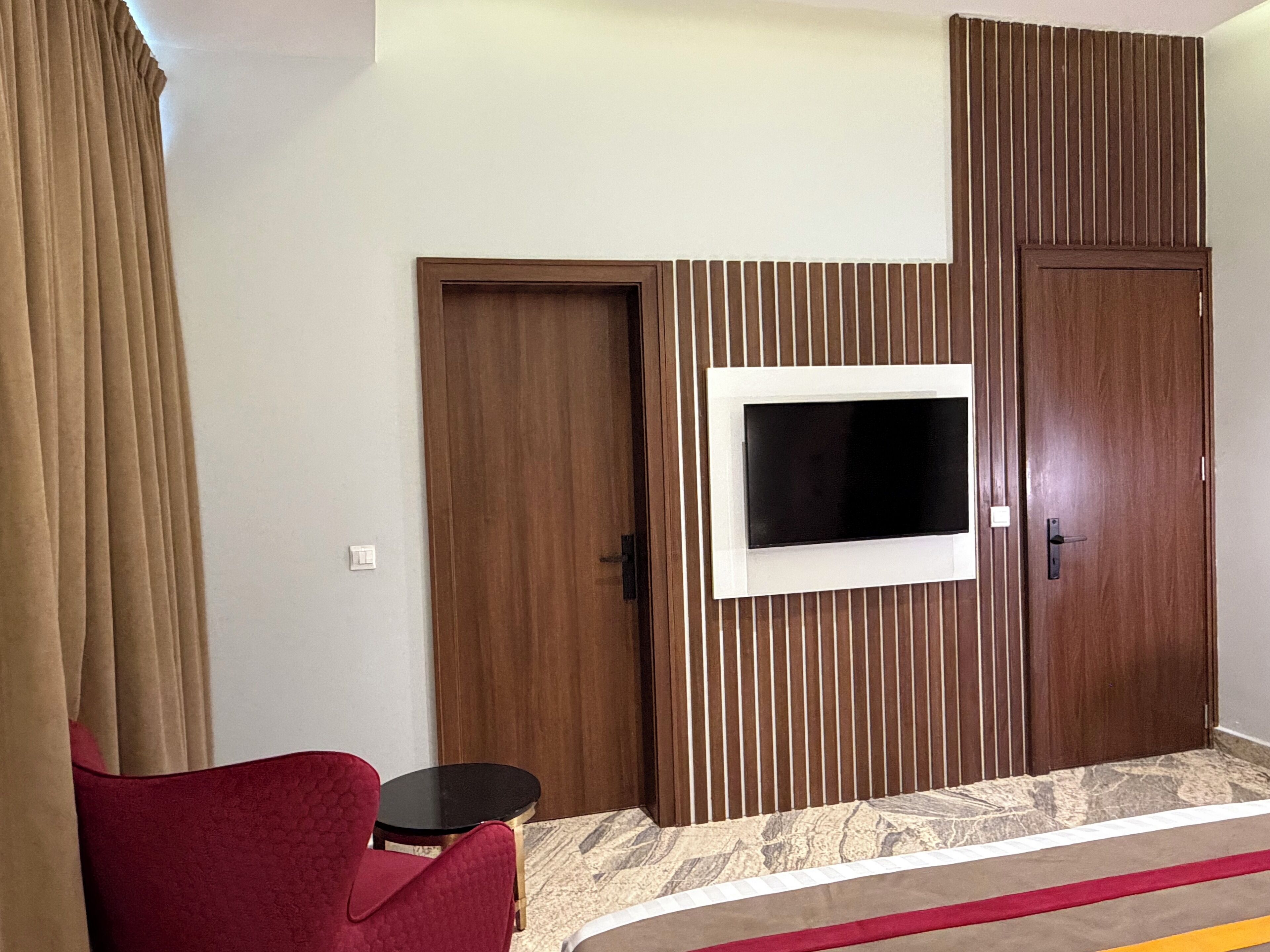 A hotel room with a wooden wardrobe, a television mounted on the wall, a small round table, and a red chair.