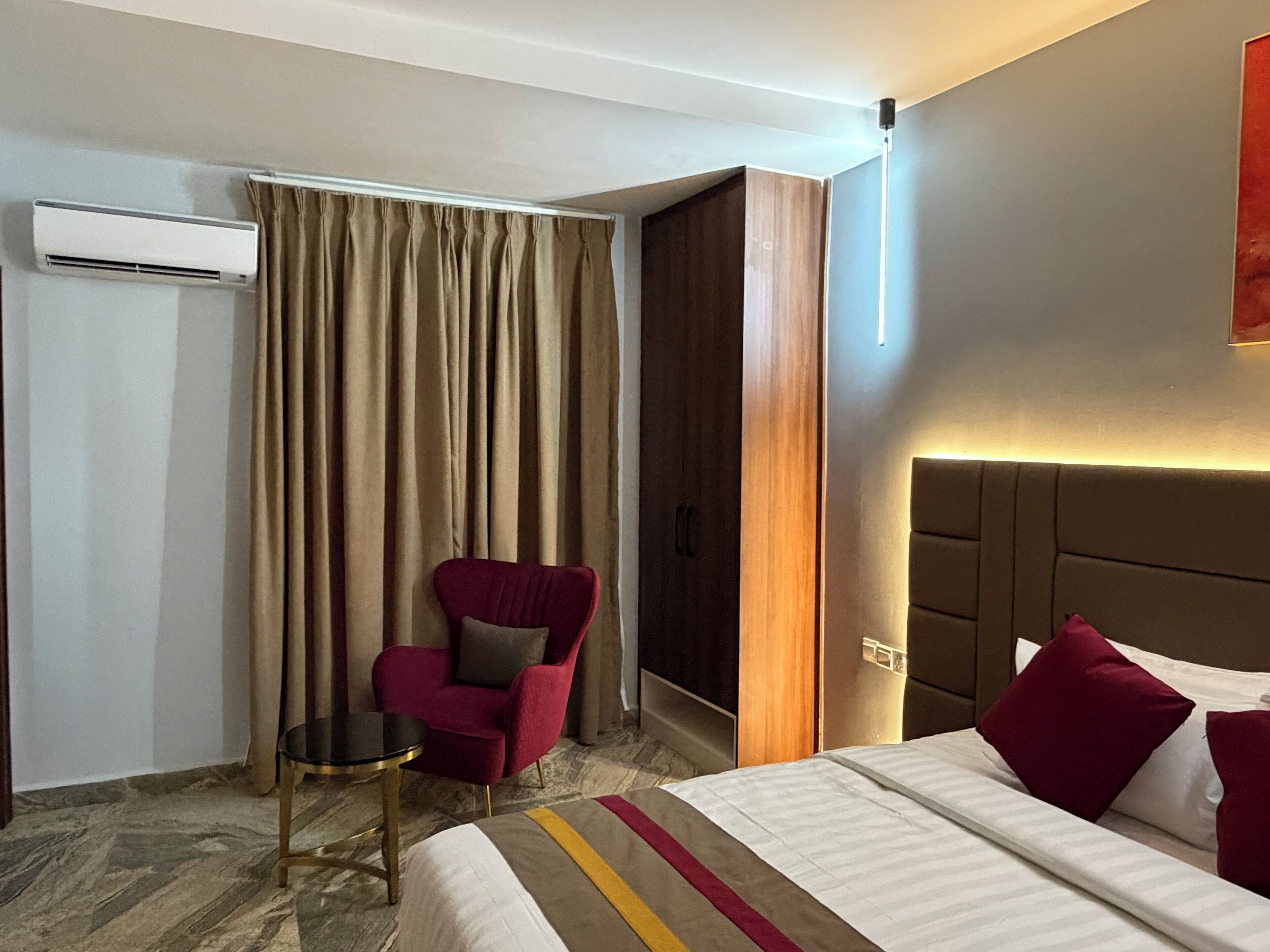 A hotel room with a bed, a red armchair, a small table, a wardrobe, and an air conditioning unit.
