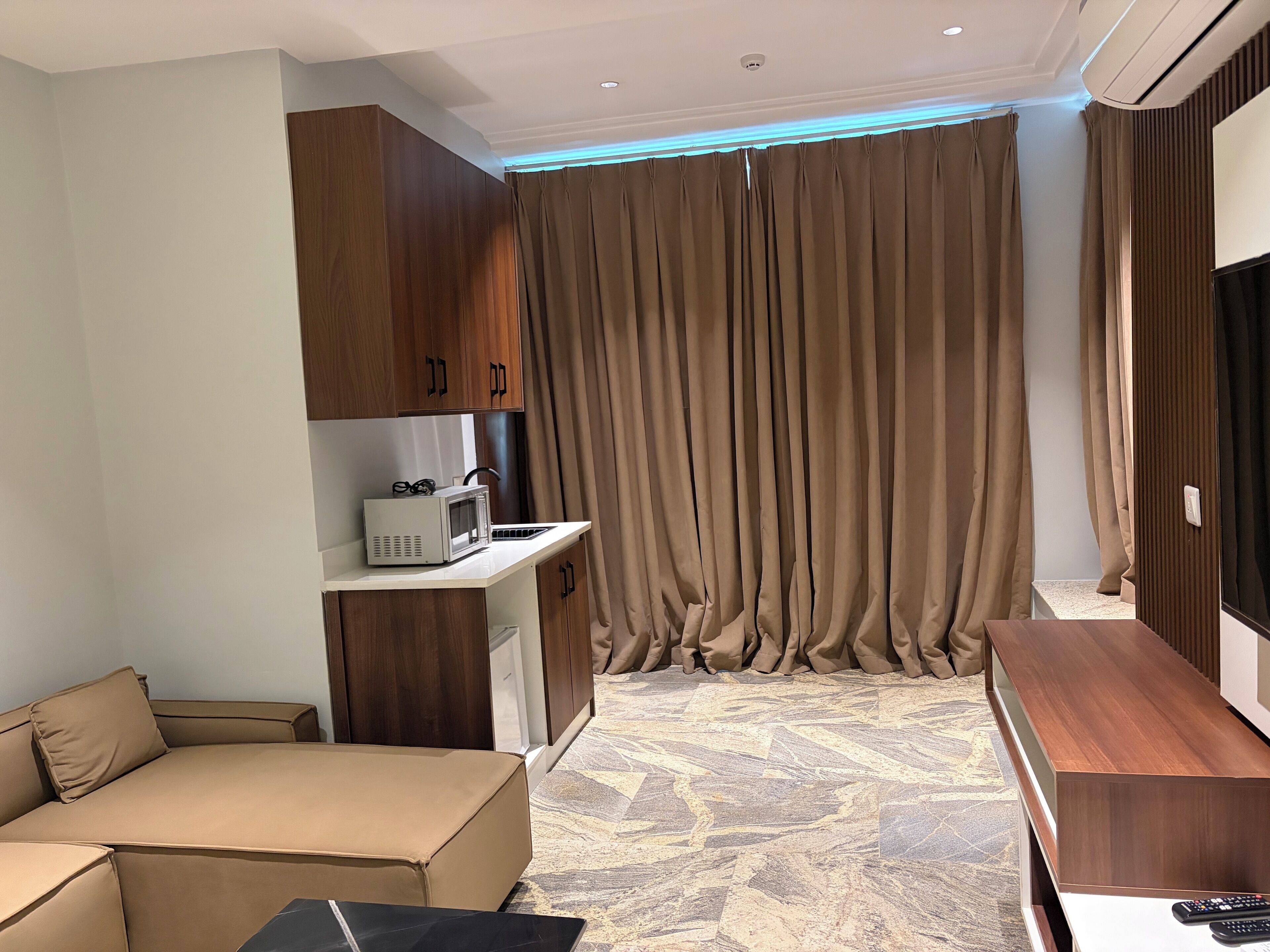 Une chambre d’hôtel moderne équipée d’un canapé, d’une petite kitchenette, d’une télévision et d’une grande fenêtre avec des rideaux.
