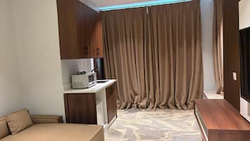 Habitación de hotel moderna con sofá, una kitchenette pequeña, televisión y un ventanal con cortinas.