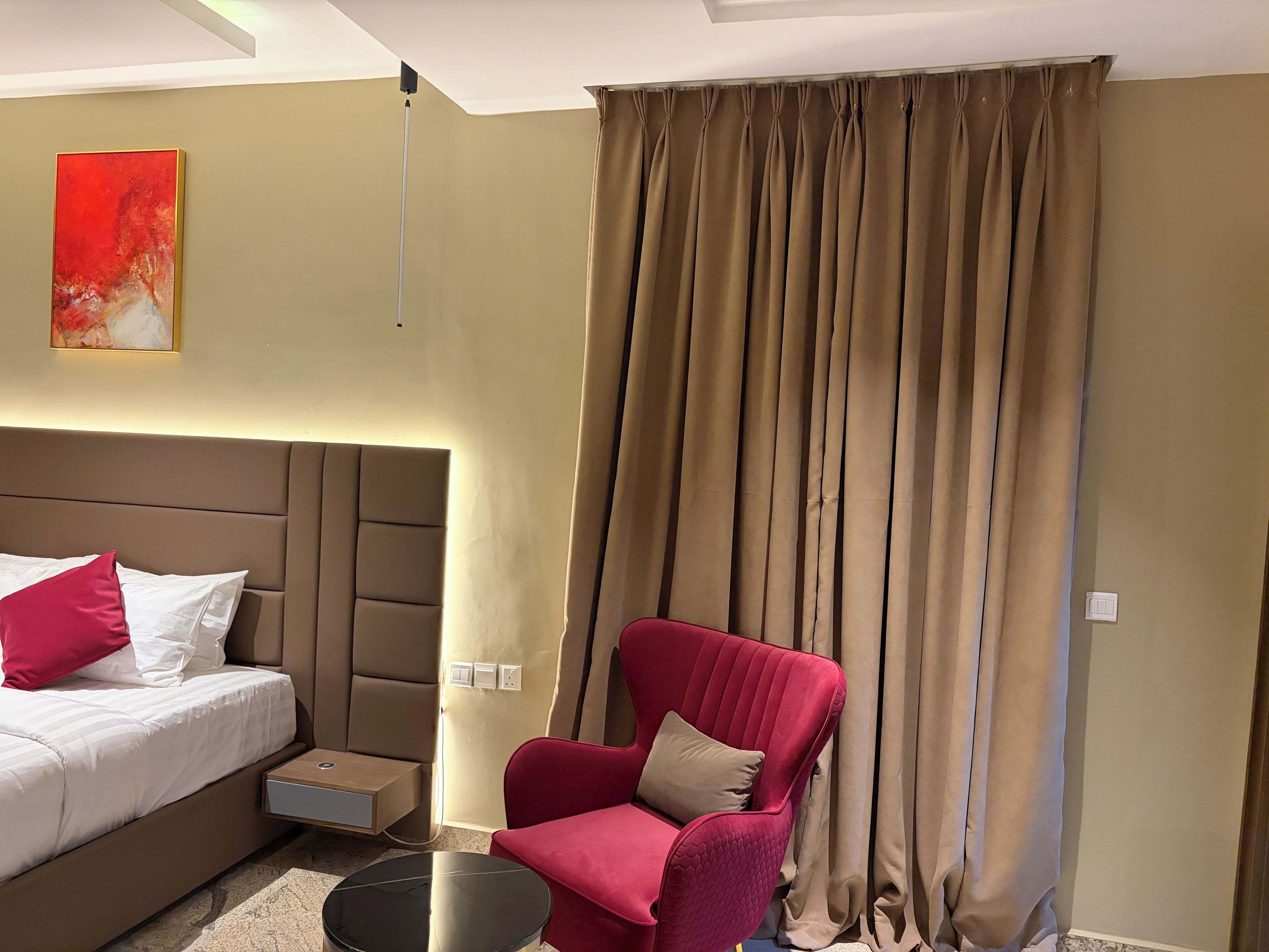 A hotel room with a bed, a red armchair, a small table, and a large window with curtains.