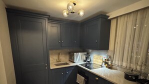 A compact kitchen with dark blue cabinets, a sink, a stove, and a window with curtains. - Soho 360 Views Flat (London)