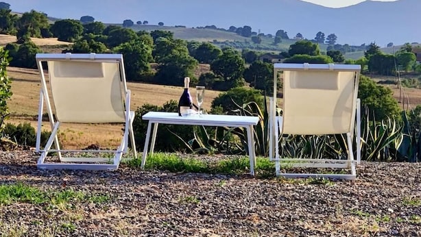 Deux fauteuils de jardin blancs, disposés de part et d’autre d’une table, sont placés sur une surface de gravier, offrant une vue sur des collines ondulantes et des arbres en arrière-plan.