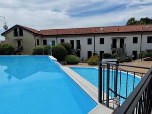 A swimming pool with a clear blue water surface, surrounded by a building with multiple windows and balconies, and a tiled roof. - App.12- Le Terrazze del Garda Immobili e Soluzioni (Manerba del Garda)