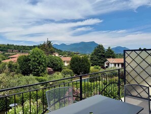 En balkon med et bord og stole, udsigt over et bjerglandskab med bygninger og grøn vegetation.