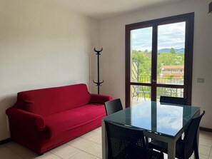 Ein Zimmer mit einem roten Sofa, einem Tisch mit Glasplatte und Stühlen, mit Blick auf Grünflächen und Gebäude durch ein großes Fenster.