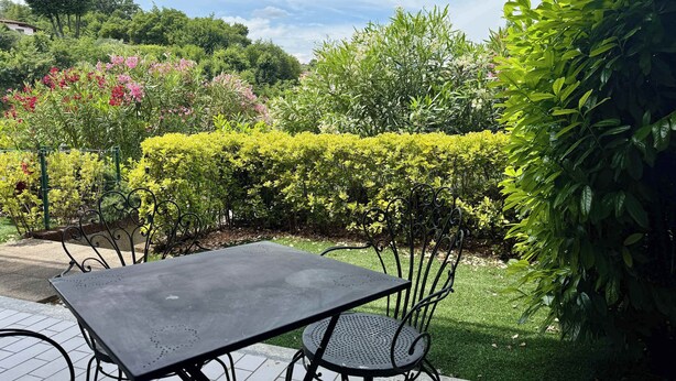 Un patio con mesa y sillas, rodeado de setos cuidados y plantas con flores.