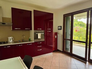 A kitchen with red cabinets, a stove, a sink, and a window with a view of greenery. - App.3- Le Terrazze del Garda Immobili e Soluzioni (Manerba del Garda)