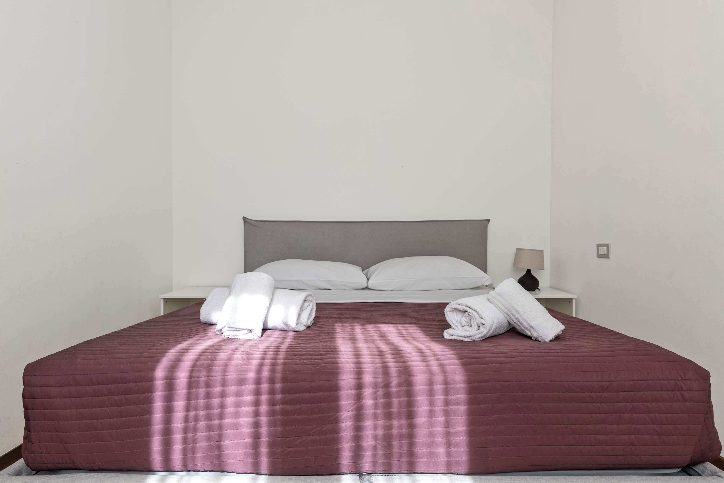 Una cama bien hecha con una colcha roja, almohadas blancas y toallas dobladas a un lado.