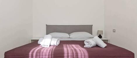 Una cama bien hecha con una colcha roja, almohadas blancas y toallas dobladas a un lado.