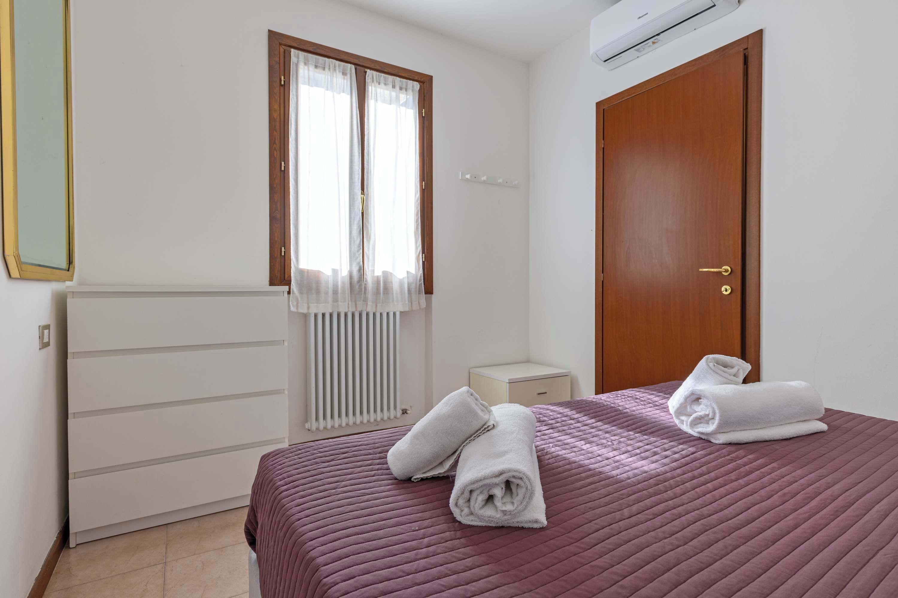 A bedroom with a bed, two towels, a window with curtains, a radiator, a wooden door, and a white bedside table.