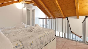 Et soveværelse med træloft, en seng med blomstret dynebetræk, en hvid radiator og en balkon med gelænder.