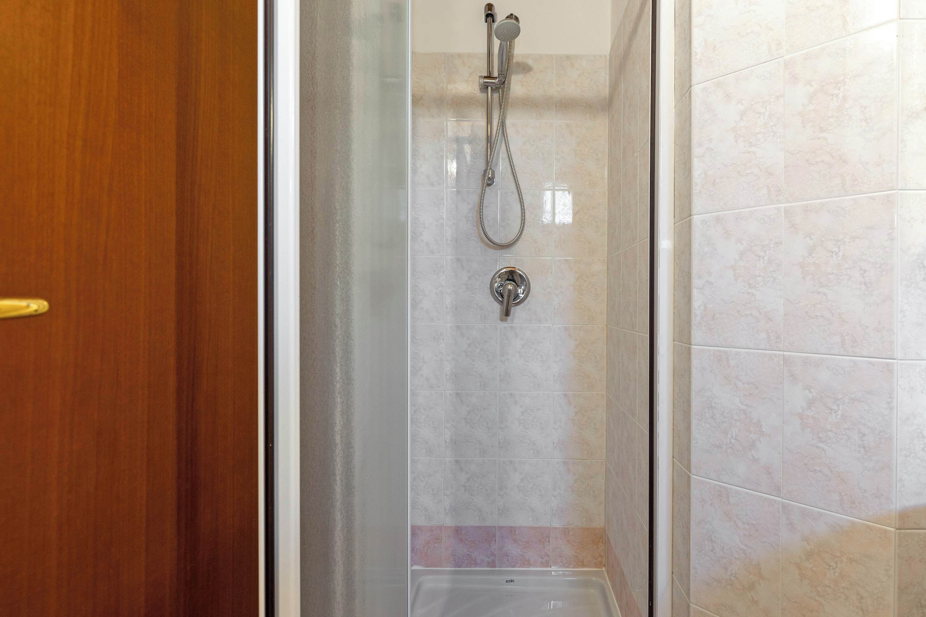 Dusche mit Handbrause, Wandarmatur, Glastür und gefliesten Wänden.