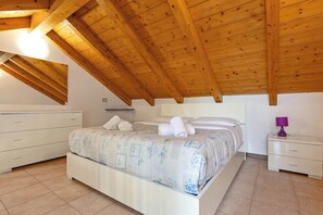 A bedroom with a wooden ceiling, a bed with a floral bedspread, a nightstand with a lamp, and a dresser. - App.17- Le Terrazze del Garda Immobili e Soluzioni (Manerba del Garda)