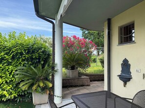 Un patio techado con mesa y sillas, rodeado de abundante follaje y plantas con flores.