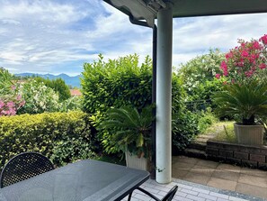 Un patio techado con mesa y sillas, rodeado de plantas diversas y vistas a montañas.