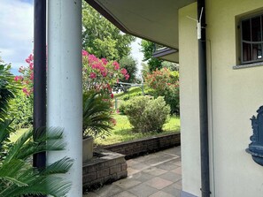 A covered outdoor area with a garden view, a stone pathway, and a building with a window and a decorative element on the wall. - App.1- Le Terrazze del Garda Immobili e Soluzioni (Manerba del Garda)