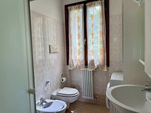 Ein Badezimmer mit Toilette, Bidet, Waschbecken und einem Fenster mit floralem Vorhang.