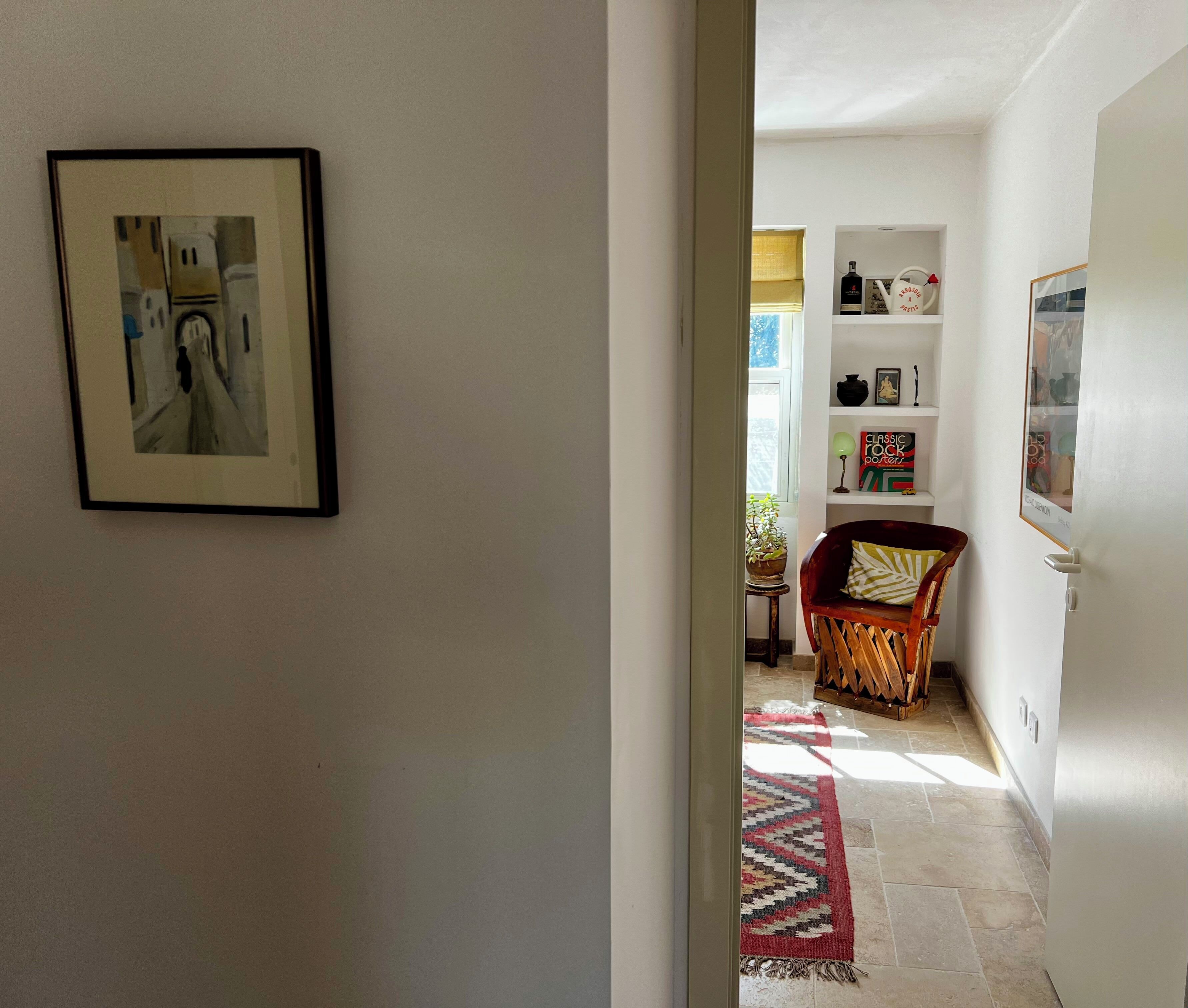 A hallway with a framed picture, a chair with a patterned cushion, a small table with a plant, and a rug on the floor.