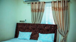A bedroom with a wooden headboard, two pillows, and beige curtains. - Superb 2-BR Duplex With 24HRS Pwr + Unlimited WiFi (Lagos)