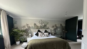 A bedroom with a large bed, a nightstand, a lamp, a potted plant, and a mural on the wall.