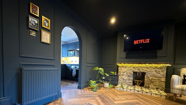 A modern living room with a fireplace, a flat-screen TV displaying 'NETFLIX', and a kitchen visible through an archway.