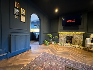 Una sala de estar moderna con chimenea, un televisor de pantalla plana mostrando 'NETFLIX', y una cocina visible a través de un arco.