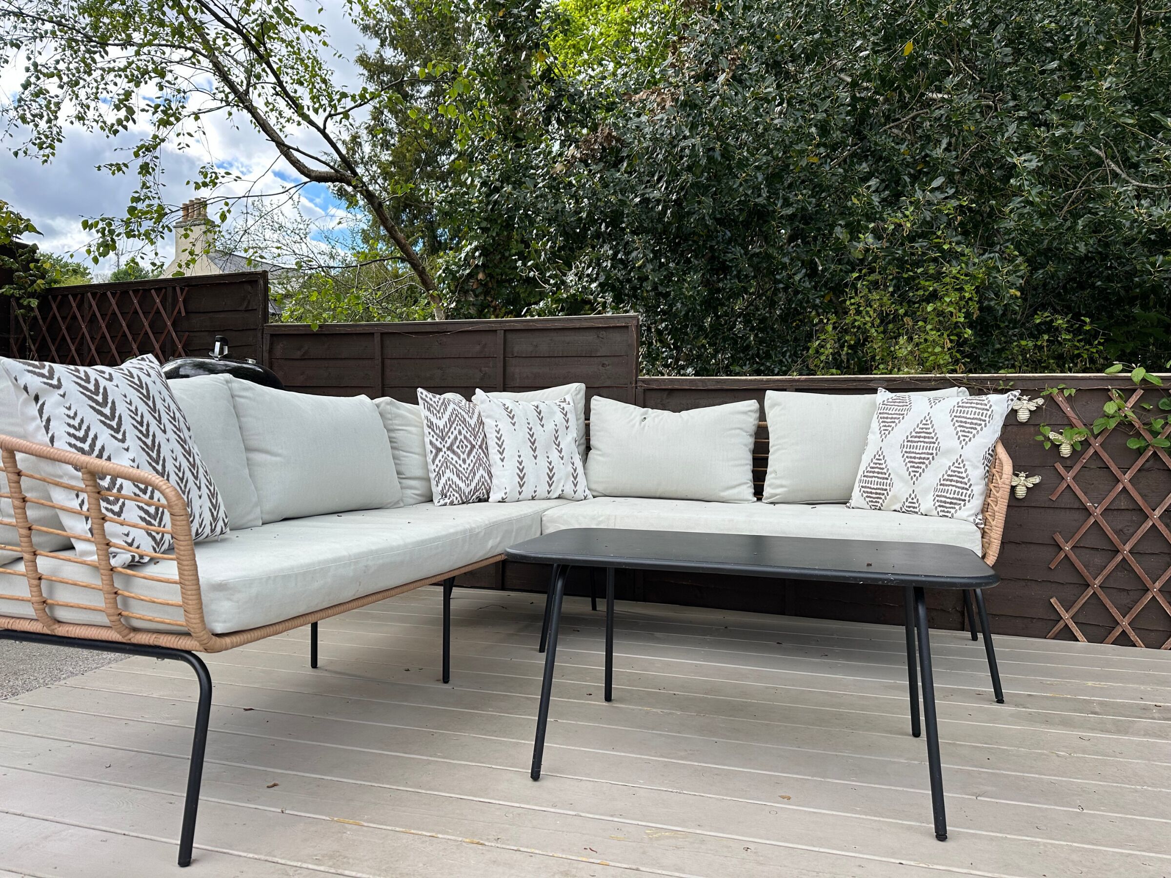 Un sofá modular exterior con cojines estampados sobre una terraza de madera, rodeado de follaje denso y una valla de madera.