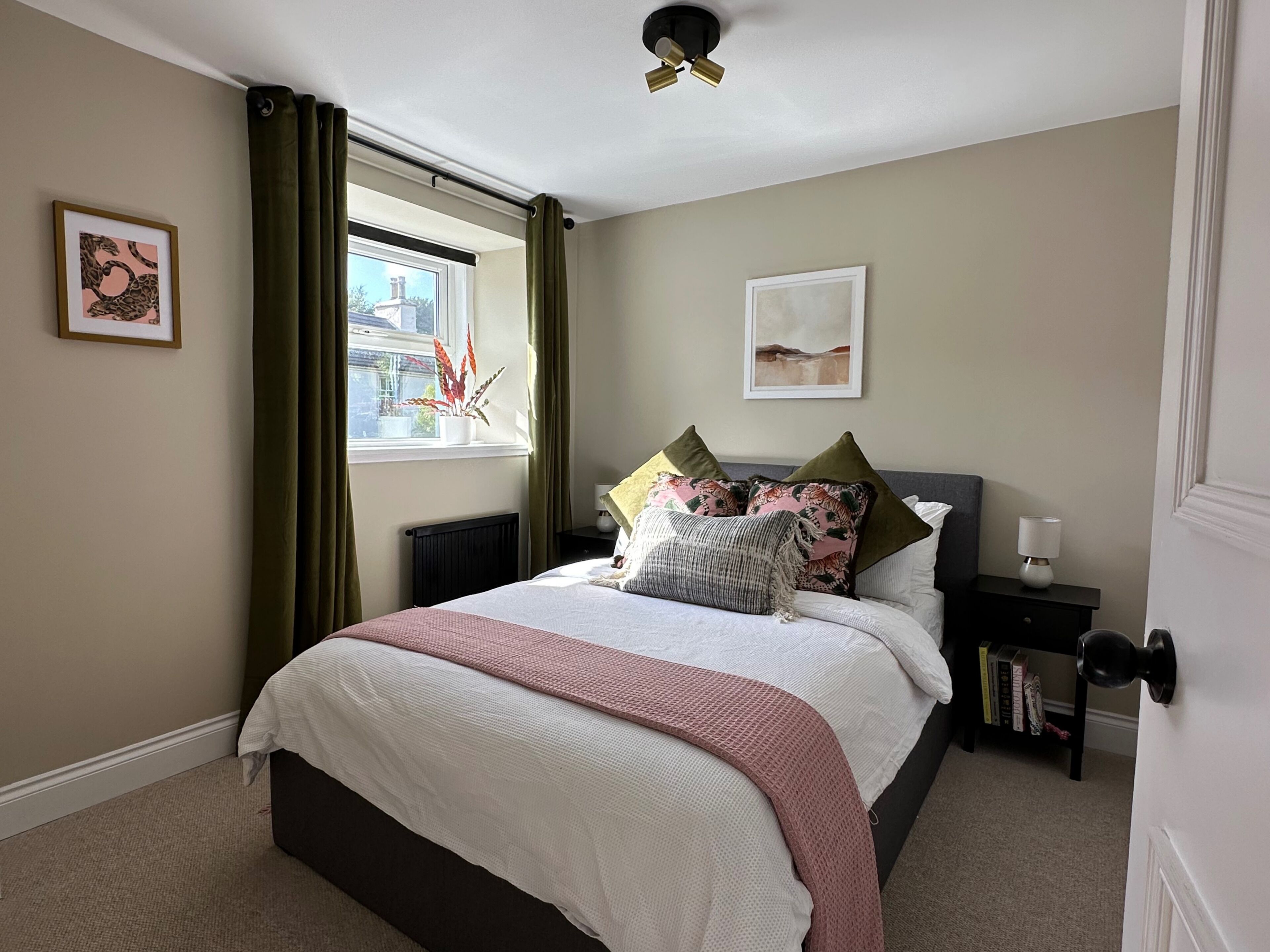 A bedroom with a bed, a window with curtains, a radiator, a bedside table with books, and a wall with framed artwork.