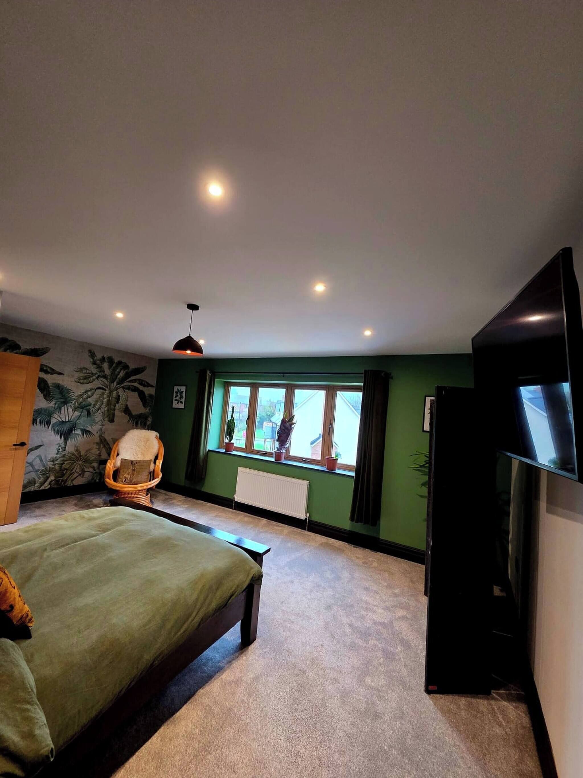 Una habitación con cama, una silla, un radiador, una ventana con cortinas y un televisor.
