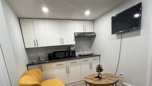 Una cocina funcional con armarios blancos, microondas y cafetera. Hay una silla amarilla y una mesa de madera redonda con un tapete colorido. Un televisor de pantalla plana está montado en la pared.
