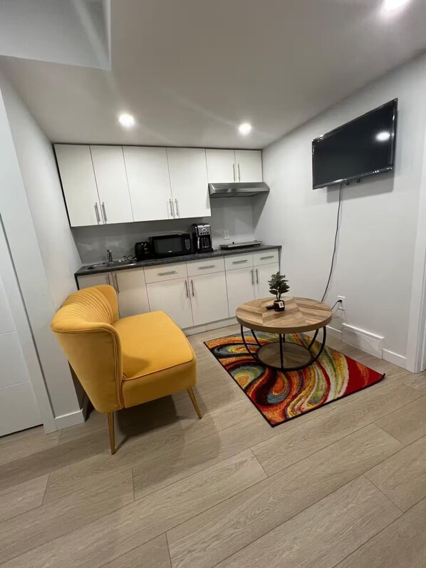 A compact kitchen with white cabinets, a microwave, and a coffee maker. There's a yellow chair and a round wooden table with a colorful rug. A flat-screen TV is mounted on the wall. - Base rate includes1bedroom (up to 2guest)need both bedroom book double base rate (Winnipeg)